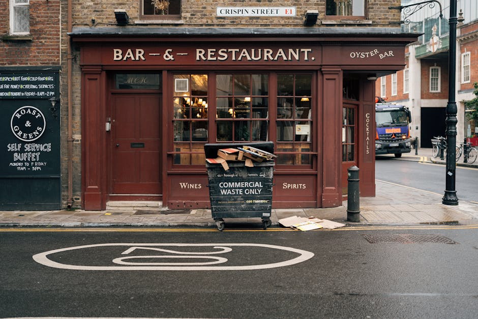 A black wheeled waste bin marked 'Commercial Waste Only' is positioned on the sidewalk in front of a brick-walled restaurant with large glass windows and a wooden exterior painted in a deep reddish-brown color. The bin contains various flattened cardboard boxes and loose paper waste, some of which are spilling onto the pavement nearby. The restaurant signage indicates it is a bar and restaurant serving wines, spirits, and cocktails, with a small blackboard to the left advertising self-service buffet hours. The scene is set on a city street, with a designated no-parking zone marked by a white circular road marking painted on the tarmac in front of the bin. To the right, a black bollard and a lamppost are visible, along with a parked truck and bicycles further down the street in the background. Bright natural lighting suggests early evening or late afternoon, and the environment appears clean but shows signs of waste collection activity typical of independent waste handling services such as those provided by Waste Collection Acton.