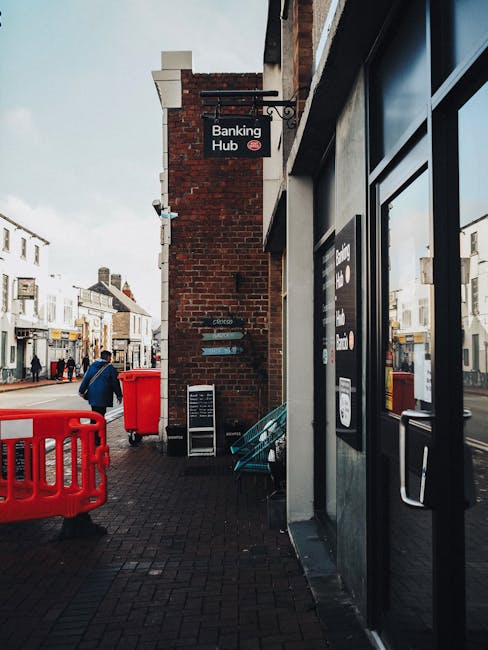 The image shows a street scene outside a building with a shop front on the right, featuring large glass doors and windows with dark frames. To the left, there is a brick wall with a sign reading 'Banking Hub' mounted on it, indicating a financial services location. On the pavement, a red waste bin and a smaller red container are visible alongside a metal barricade, which may serve as a safety barrier during street works or events. A person wearing a blue jacket and carrying a bag walks away from the building, heading along the busy street that features various shops and commercial establishments with display windows and signage on both sides. The environment appears to be an urban setting with overcast lighting, and the scene captures a typical day with pedestrians and street furniture. The setup suggests typical urban waste collection practices, with Waste Collection Acton potentially providing alternative rubbish removal or on-site clearance services for businesses in this commercial area, especially given the presence of waste containers outdoors to facilitate easy rubbish removal.