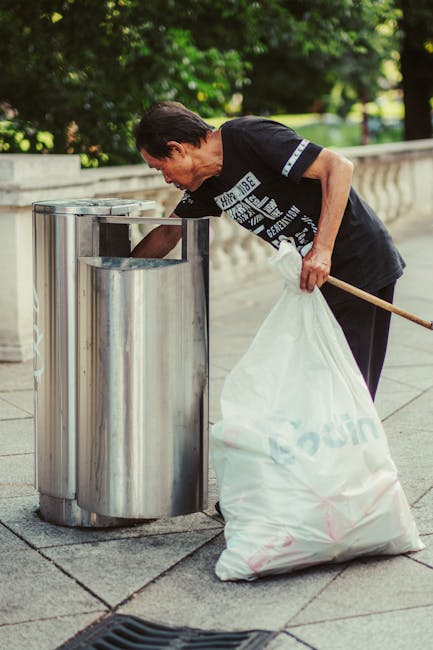 A middle-aged man with dark hair, wearing a black T-shirt with white text, is seen disposing of rubbish into a cylindrical public litter bin made of shiny brushed metal on a paved sidewalk. He holds a large white garbage bag filled with waste in his left hand while leaning forward to empty contents into the bin. The scene is set outdoors, with a row of lush green trees and a stone balustrade visible in the background, indicating an urban park or promenade area. The lighting suggests daylight, casting soft shadows on the ground. The environment appears clean and well-maintained, reflecting responsible waste disposal and the importance of litter management, which can relate to private or community rubbish collection services like those offered by Waste Collection Acton. This scene exemplifies community efforts or independent waste handling related to rubbish removal within a neighbourhood setting, highlighting proper disposal practices without the use of recycling or disposal machinery in view.